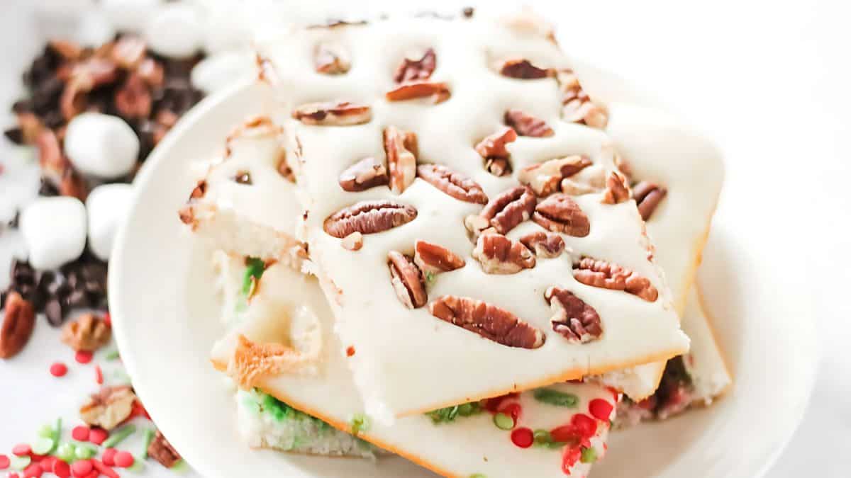 A plate of white chocolate bark topped with pecans and festive sprinkles, with marshmallows, chocolate chips, and pecans scattered in the background.