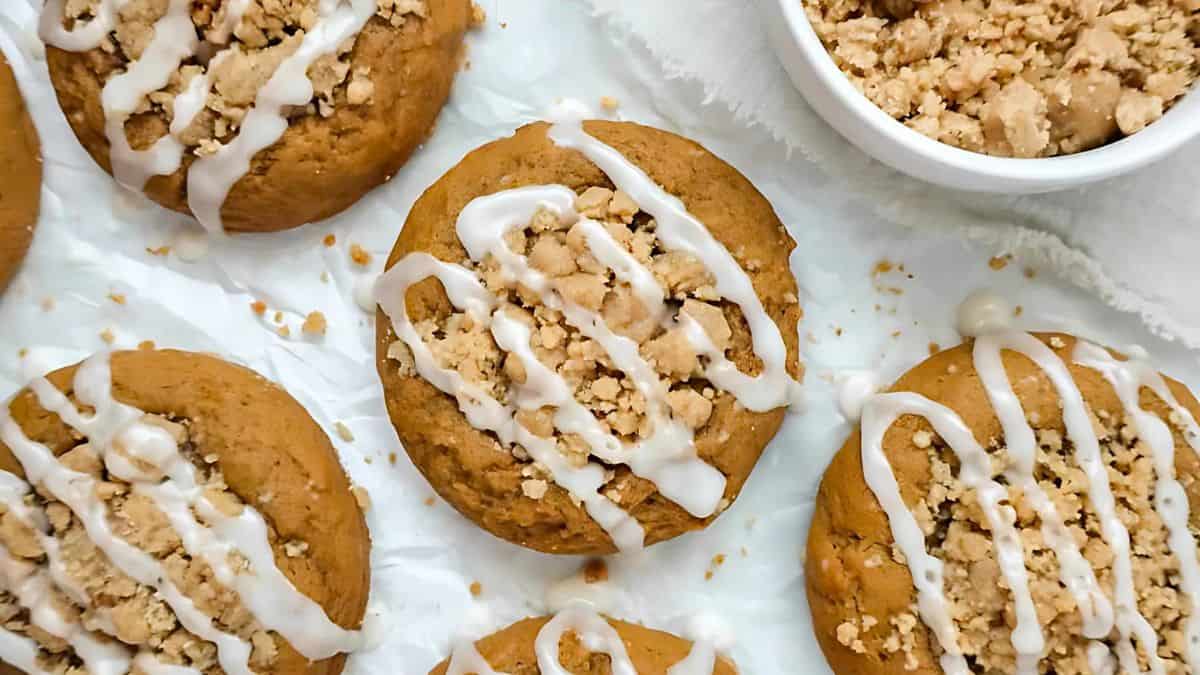 Close-up of pumpkin cookies topped with white icing drizzle and streusel, arranged on parchment paper. A small bowl of additional streusel is visible in the corner.