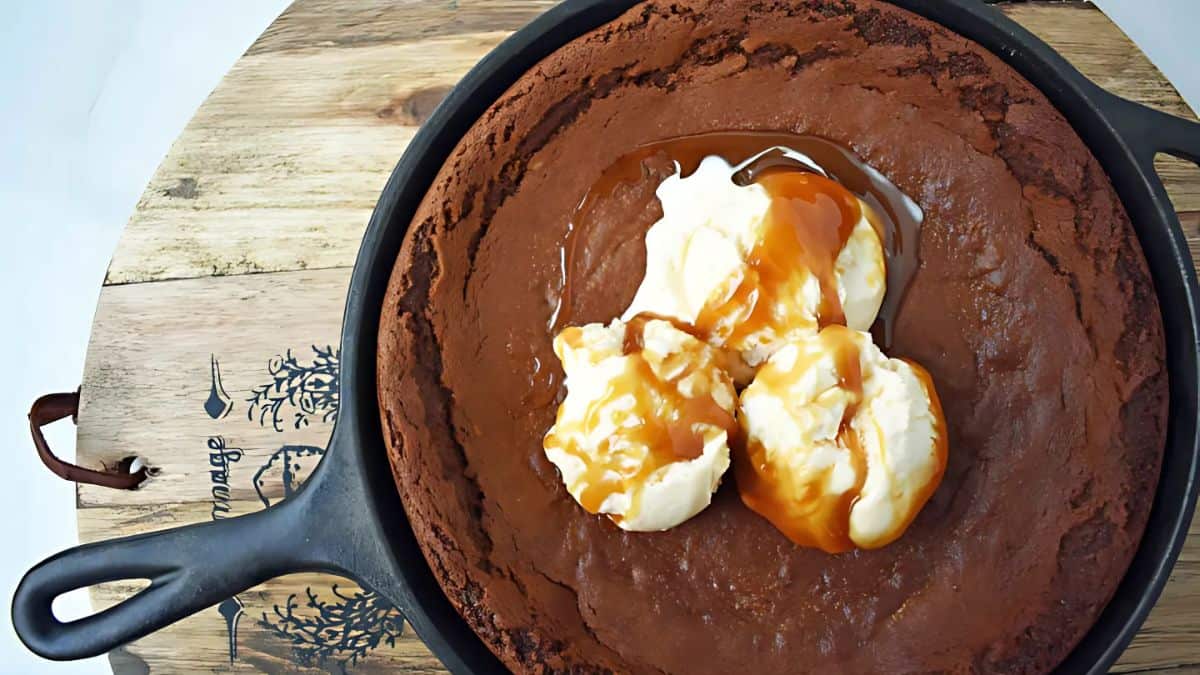 A cast iron skillet holds a large, round chocolate brownie topped with scoops of vanilla ice cream drizzled with caramel sauce. The skillet rests on a wooden board with decorative patterns.