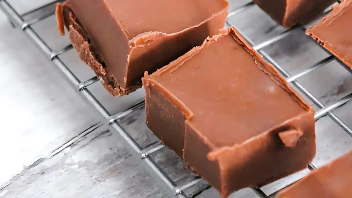 Close-up of several pieces of chocolate fudge resting on a wire cooling rack. The fudge has a smooth, glossy surface and sharp edges, indicating precise cuts. The background is a textured light gray surface.