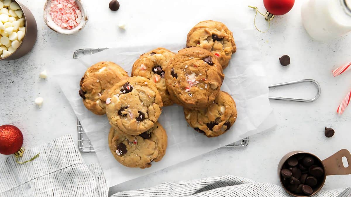A batch of freshly baked chocolate chip cookies is arranged on parchment paper on a cooling rack. Next to the cookies are chocolate chips, a red ornament, a candy cane, and a glass of milk on a white surface.