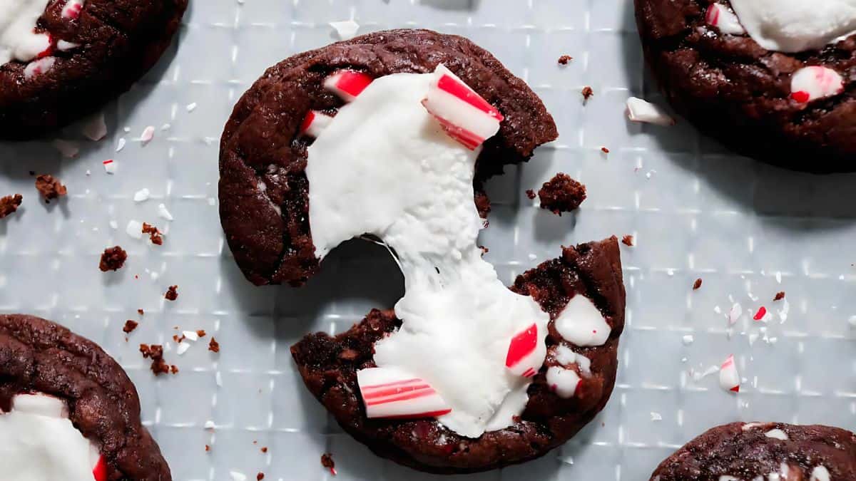 A chocolate cookie with a white filling is broken in half, revealing a gooey center. Crushed red and white peppermint pieces are scattered on top. Other similar cookies are partially visible on a grid-patterned surface.