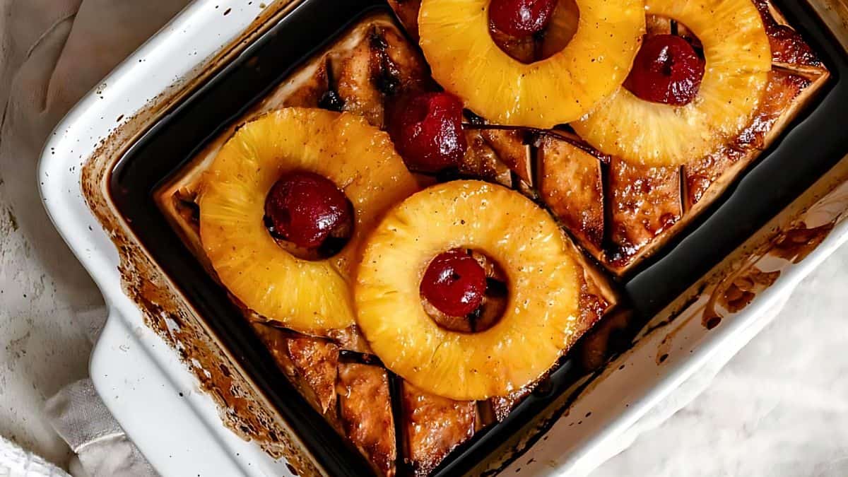 A baked dish featuring slices of glazed ham topped with pineapple rings and cherries in a rectangular pan, with a slightly browned exterior and a white cloth beside the pan.