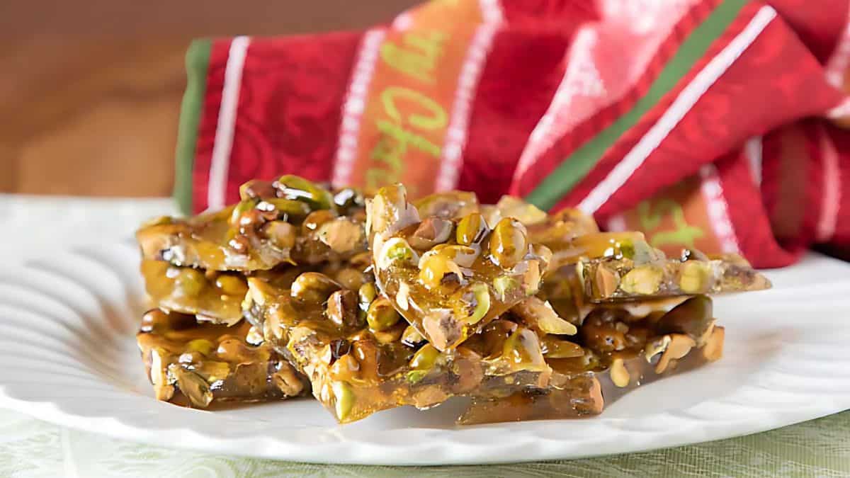 A white plate filled with pieces of nut brittle featuring a mix of pistachios and other nuts. The background includes a festive red and green cloth, adding a holiday theme to the setting.