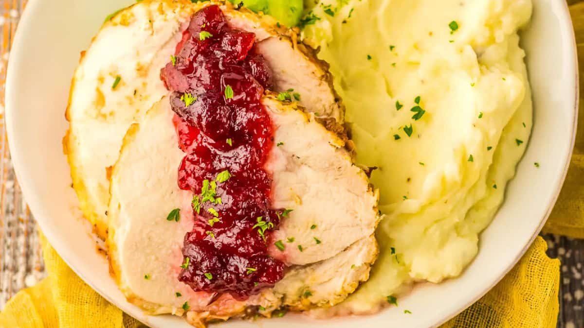 A plate featuring slices of roasted turkey breast topped with cranberry sauce, beside creamy mashed potatoes garnished with parsley. The meal is served on a white plate with a yellow napkin beneath it.
