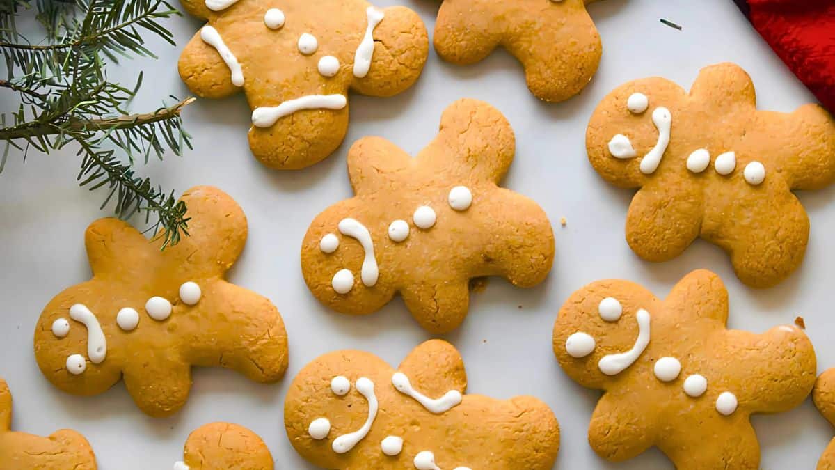A collection of gingerbread cookies shaped like people, decorated with white icing for details such as eyes, mouths, and buttons. A sprig of evergreen is visible on the left side, adding a festive touch.