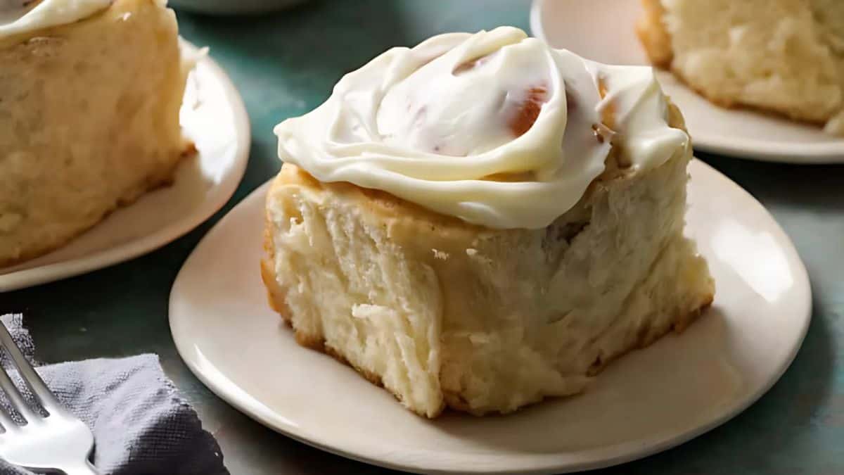 A frosted cinnamon roll sits on a white plate. The soft, fluffy dough is topped with a layer of creamy icing, with two other rolls visible in the background on similar plates. A fork rests on a napkin beside the plate.