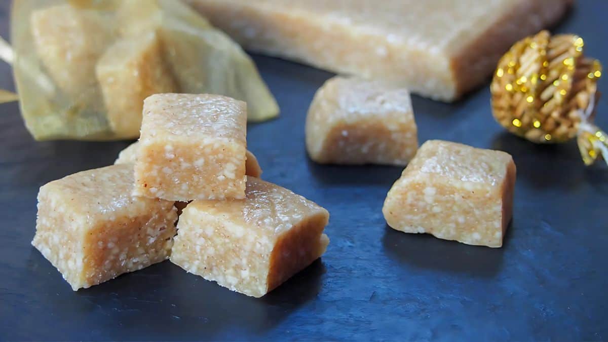 Pieces of brown, nutty candy are arranged on a dark surface. Some are stacked, and others are scattered. A large block of the same candy and a decorative golden item are visible in the background.