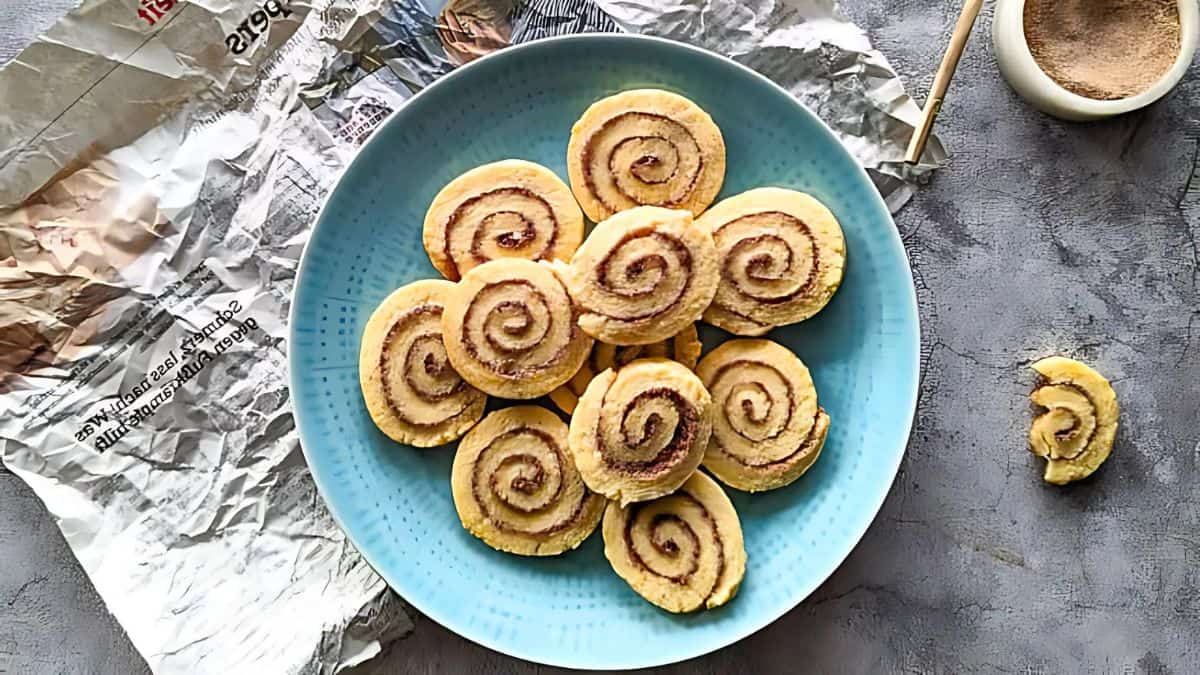 A blue plate holds a circular arrangement of cinnamon roll cookies with swirled patterns. Beside it, there is foil and a small bowl with cinnamon powder on a textured gray surface. One cookie is partially broken on the right.