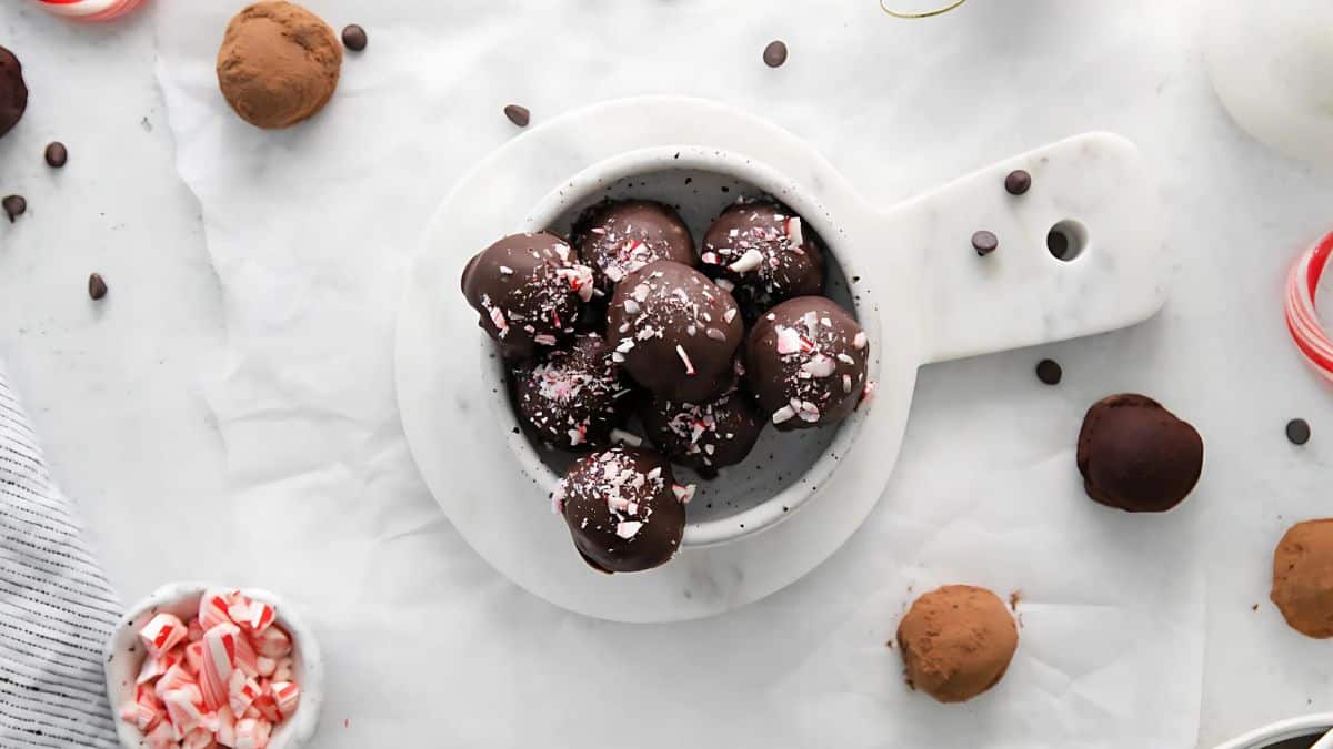 Top view of chocolate-covered truffles in a white dish, sprinkled with crushed peppermint. Additional truffles and scattered peppermint pieces are on a white surface, along with cocoa powder and chocolate chips. A striped cloth is partially visible on the side.