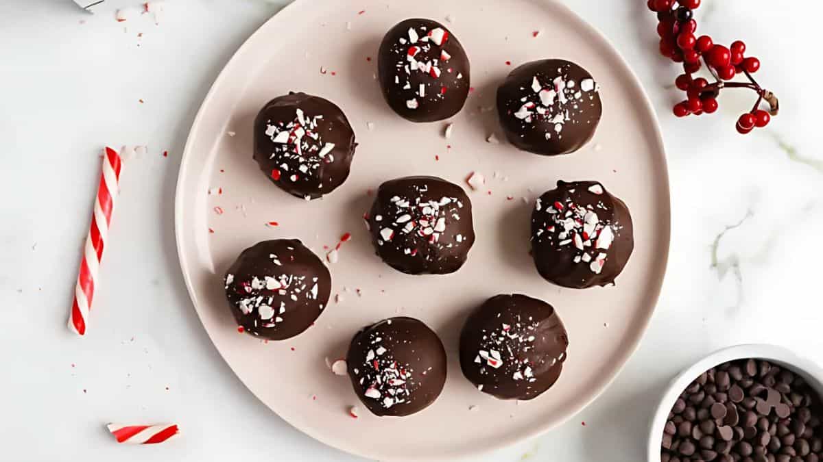 A plate of chocolate-covered truffles topped with crushed peppermint pieces. Nearby are a small bowl of chocolate chips, a sprig of red berries, and a broken candy cane on a marble surface.