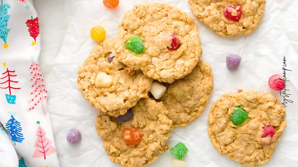 A top view of oatmeal cookies with colorful gumdrops on a crumpled sheet of parchment paper. A festive cloth with red, blue, and teal Christmas tree designs is partially visible on the left.