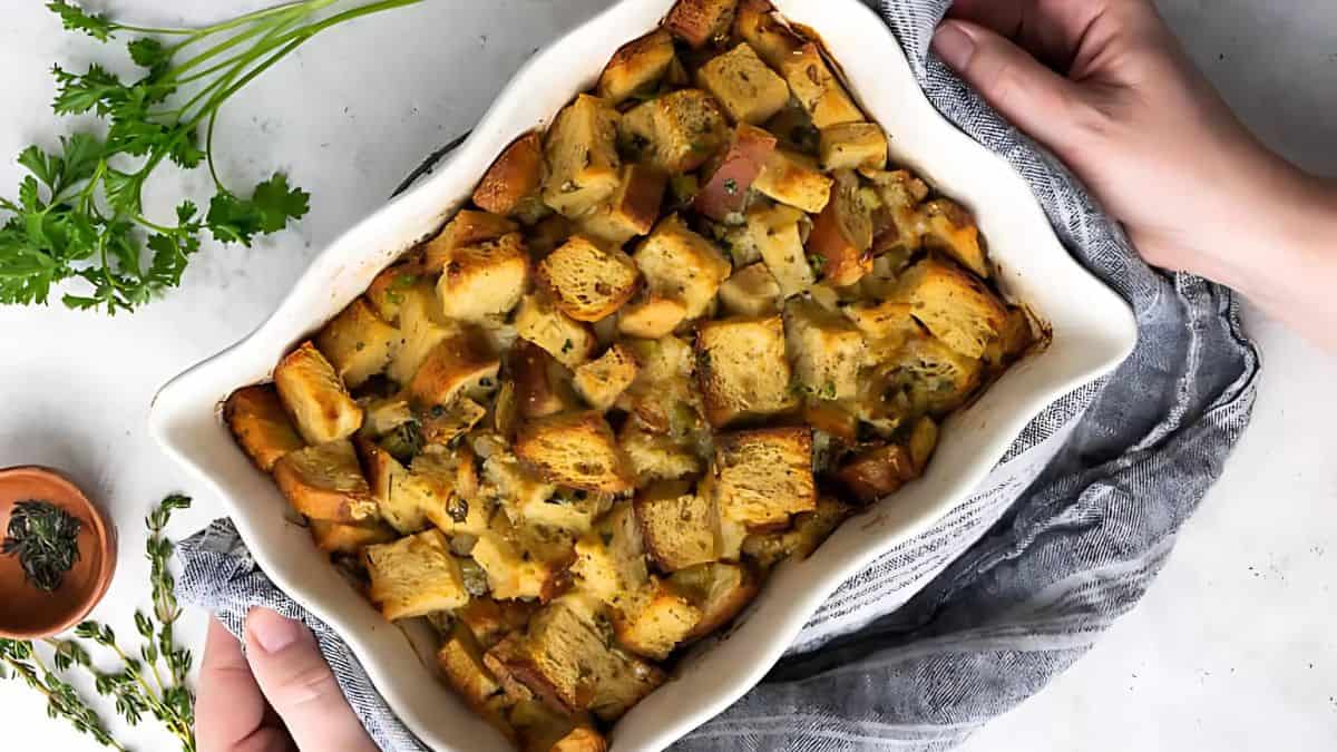 Hands holding a ceramic dish filled with baked bread stuffing, garnished with herbs. Fresh parsley and a small bowl with thyme are on the side. The dish rests on a gray cloth.