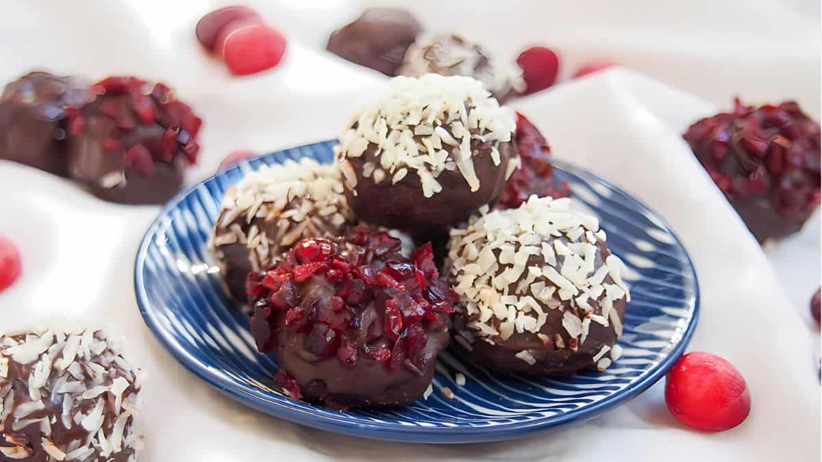 A blue plate with chocolate truffles topped with shredded coconut and chopped red fruit, set on a white cloth. Additional truffles are scattered around the plate, surrounded by a few whole red fruits.