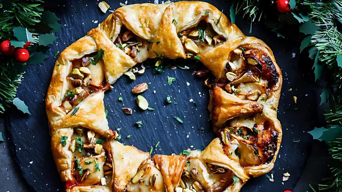 A festive puff pastry wreath filled with melted cheese and scattered with chopped nuts and herbs. It's placed on a dark serving platter, surrounded by sprigs of green foliage and red berries, creating a holiday theme.