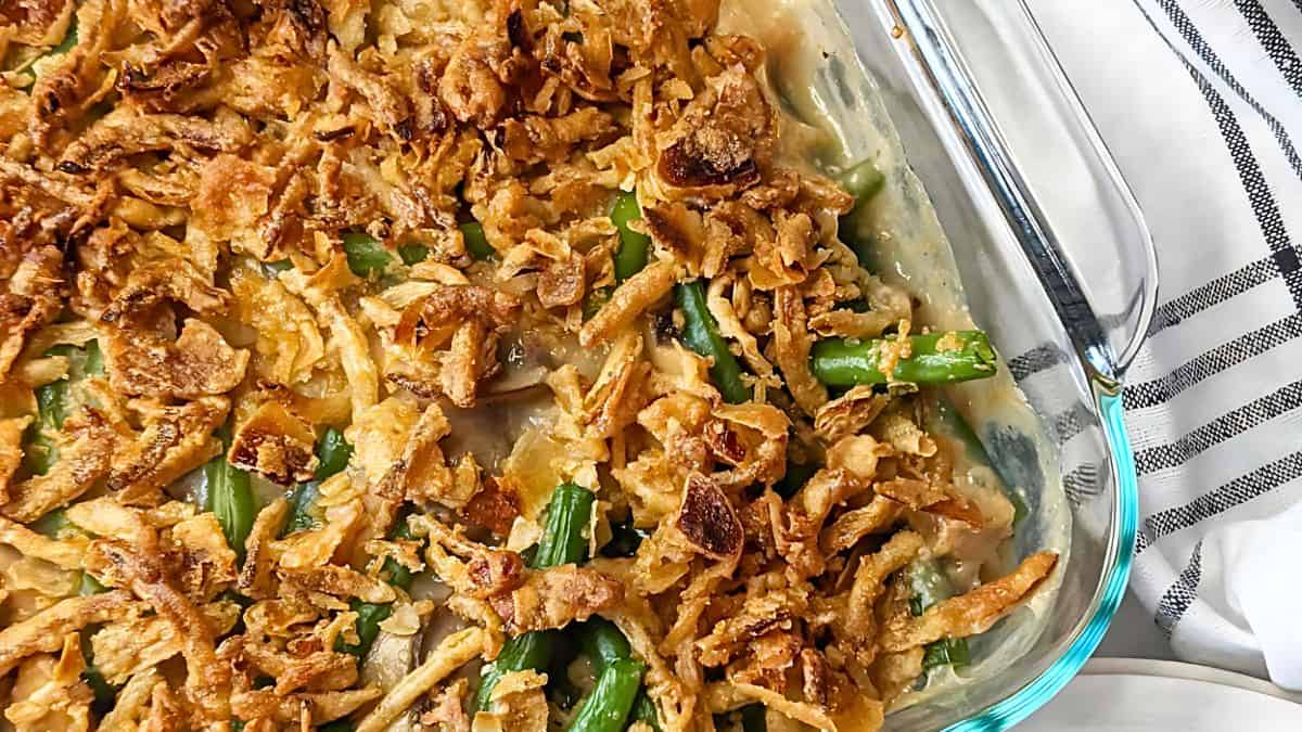 A glass baking dish filled with green bean casserole topped with crispy fried onions. The casserole is creamy and the green beans are visible through the topping. A striped cloth is partially seen on the side.