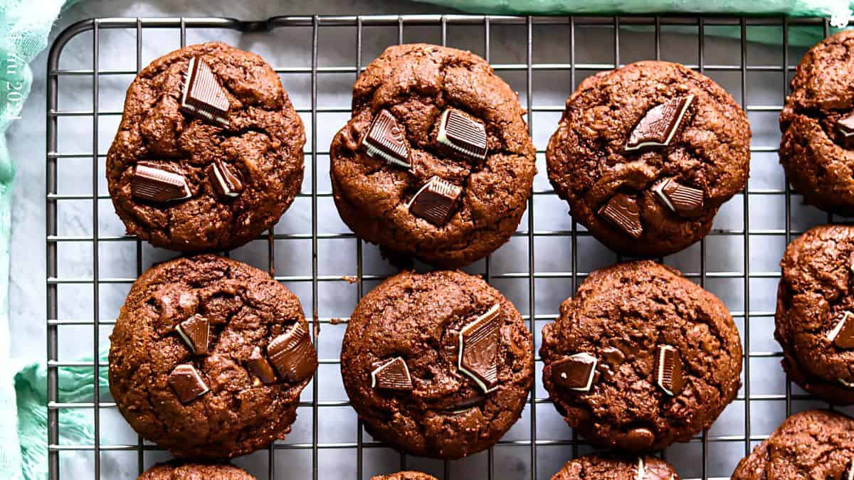 Chocolate cookies with chunks of chocolate on top are cooling on a wire rack. The cookies are arranged in neat rows, and the surface beneath the rack is a light gray.