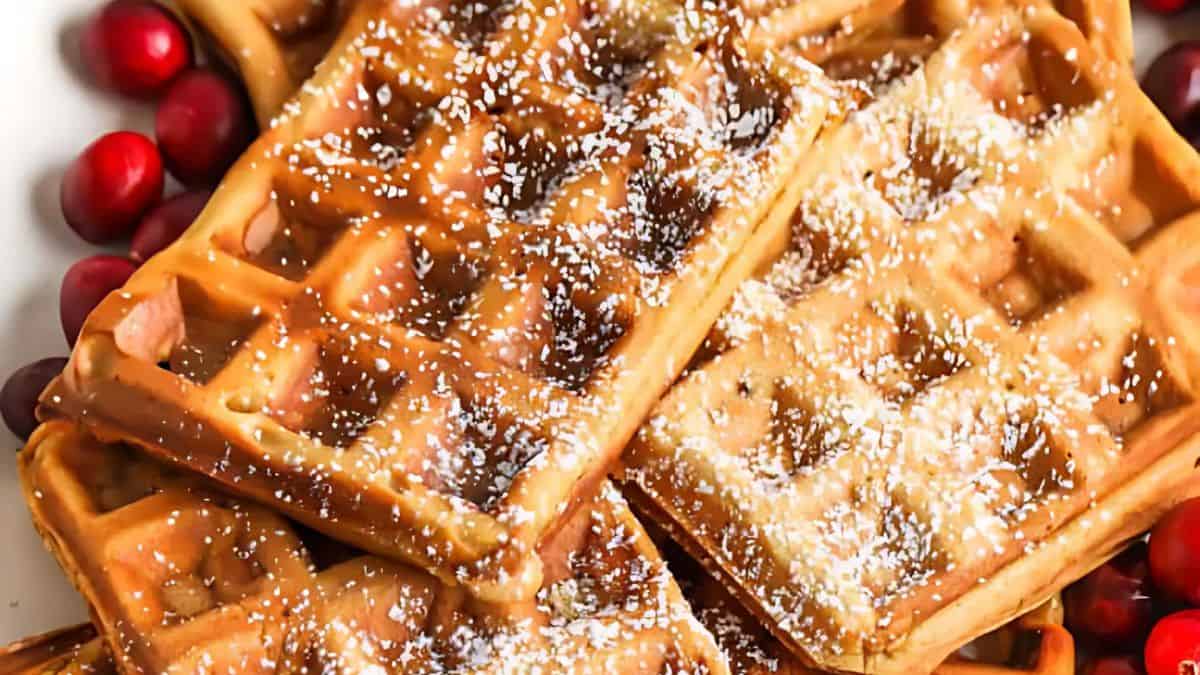A close-up of freshly made waffles sprinkled with powdered sugar, surrounded by scattered fresh cranberries. The waffles are golden brown and have a crispy texture.