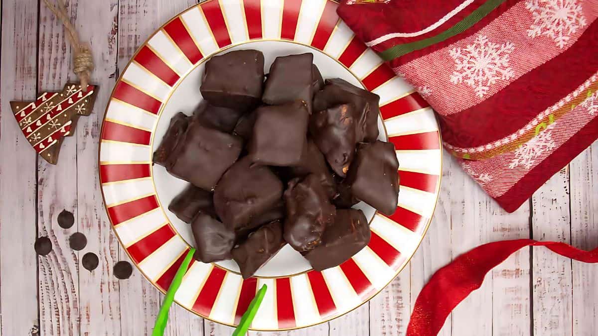 A festive plate of chocolate-covered treats is placed on a red and white striped plate. Beside it, there's a red and white snowflake-patterned cloth, a small decorated Christmas tree ornament, chocolate chips, and green-handled forks on a wooden surface.
