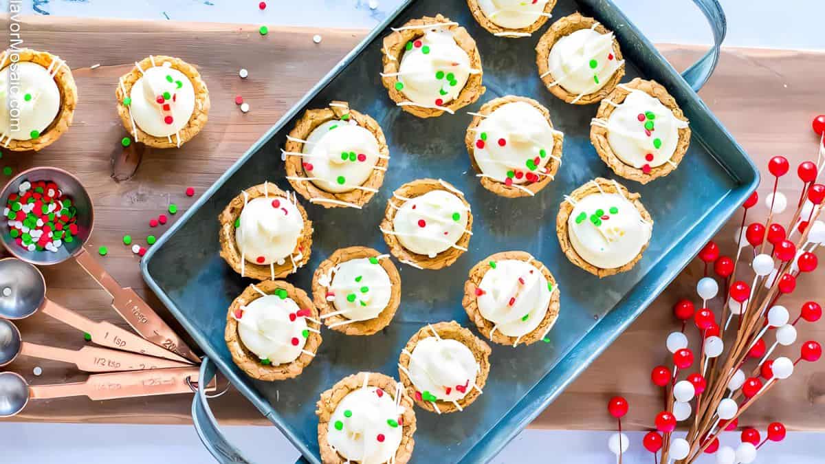 A tray of festive mini tarts topped with white frosting and sprinkled with red and green decorations. Nearby are measuring spoons and a decorative branch with red and white berries.