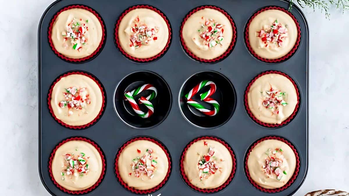 A muffin tray holding nine cupcake batter cups sprinkled with crushed candy canes. Two empty slots contain small candy canes arranged in a swirl pattern. The tray rests on a light countertop, with some greenery in the corner.