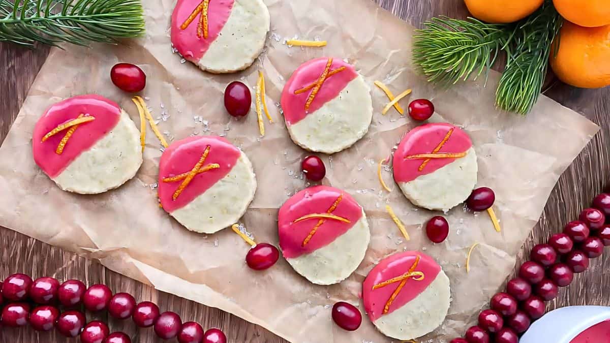 Festive cookies with pink icing and orange zest on parchment paper, surrounded by cranberries, evergreen sprigs, and oranges, creating a holiday-themed setting.