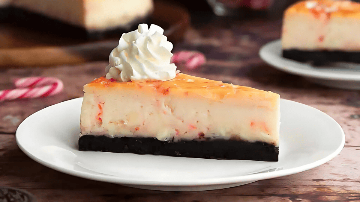 A slice of cheesecake with an orange topping on a dark crust, served on a white plate. The cheesecake is garnished with a swirl of whipped cream on top. The background features another plate and blurred red and white candies.