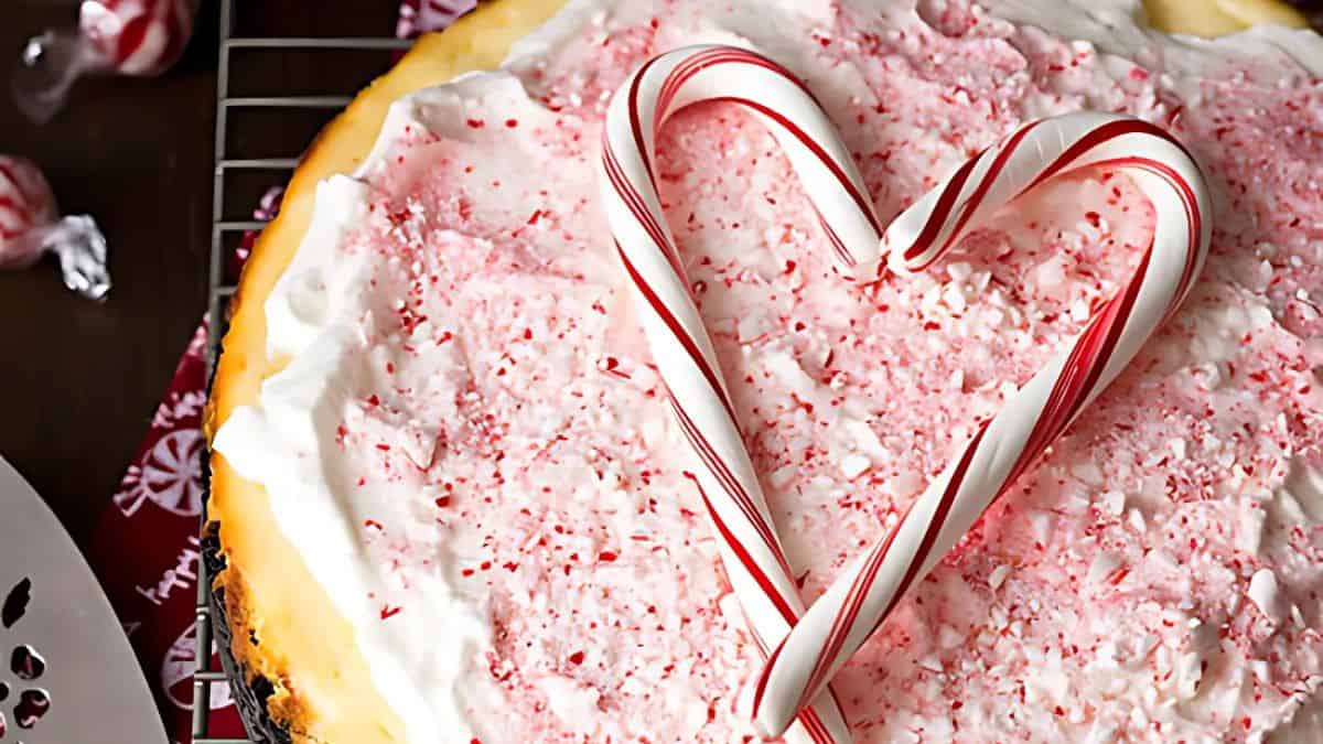 A cheesecake topped with white frosting and crushed peppermint candy, shaped like a heart using two candy canes, sits on a cooling rack. Peppermint candies are scattered around for decoration.