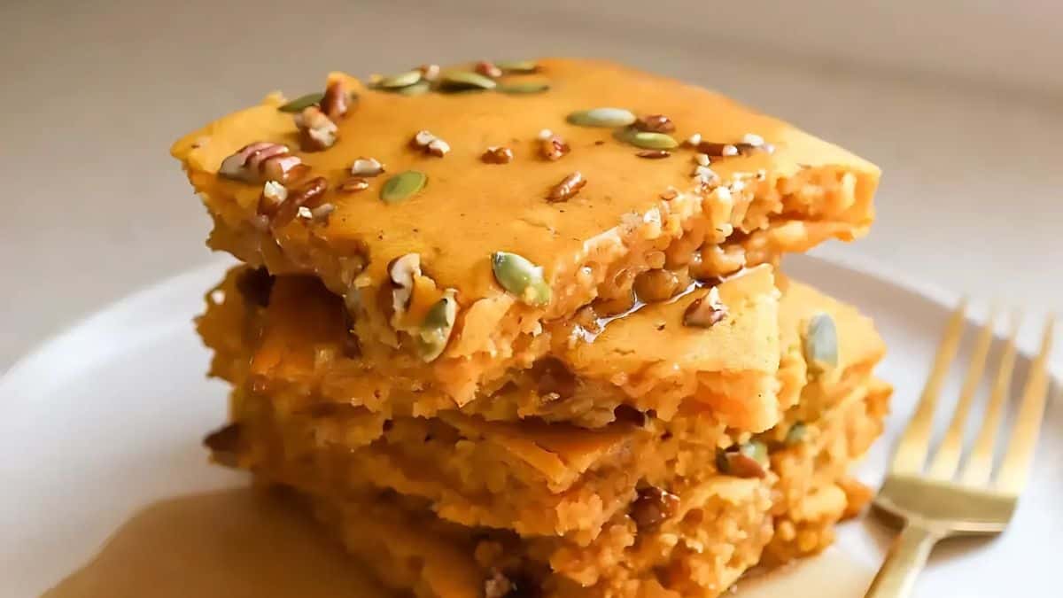 A stack of homemade pumpkin pancakes topped with pecans and pumpkin seeds, drizzled with syrup, on a white plate. A silver fork lies beside the plate.