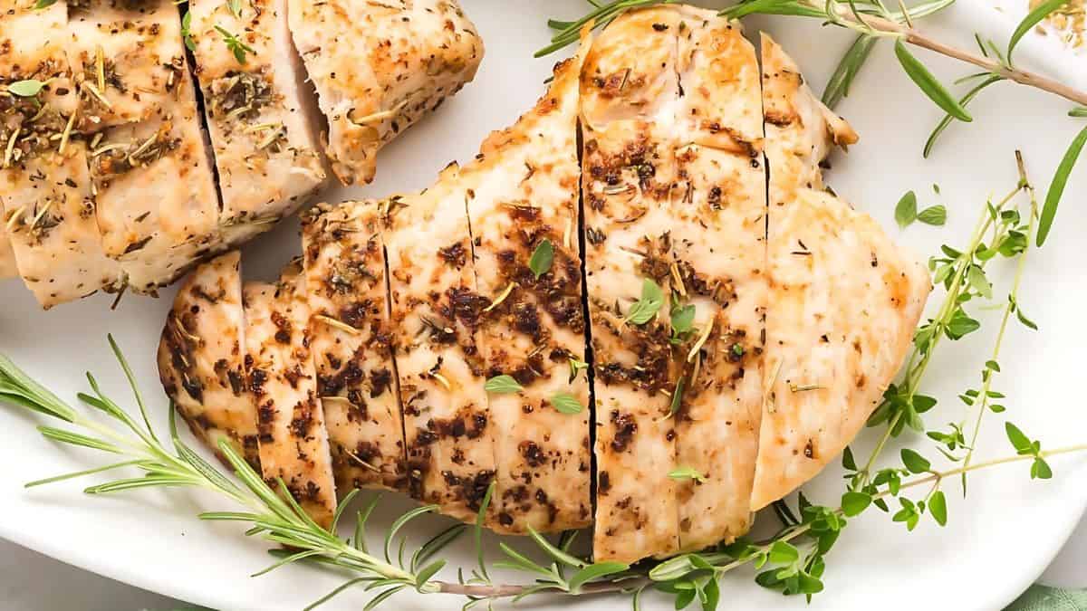 Sliced, seasoned grilled chicken breast garnished with fresh rosemary and thyme on a white plate.