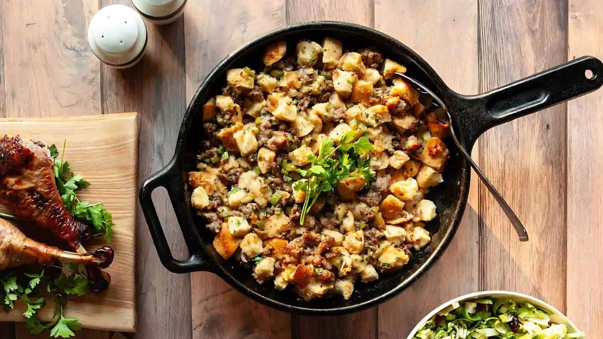 A cast-iron skillet filled with a savory stuffing mixture topped with parsley sits on a wooden table. Nearby, roasted chicken legs are on a cutting board, and a bowl of green salad is visible. Salt and pepper shakers are in the background.