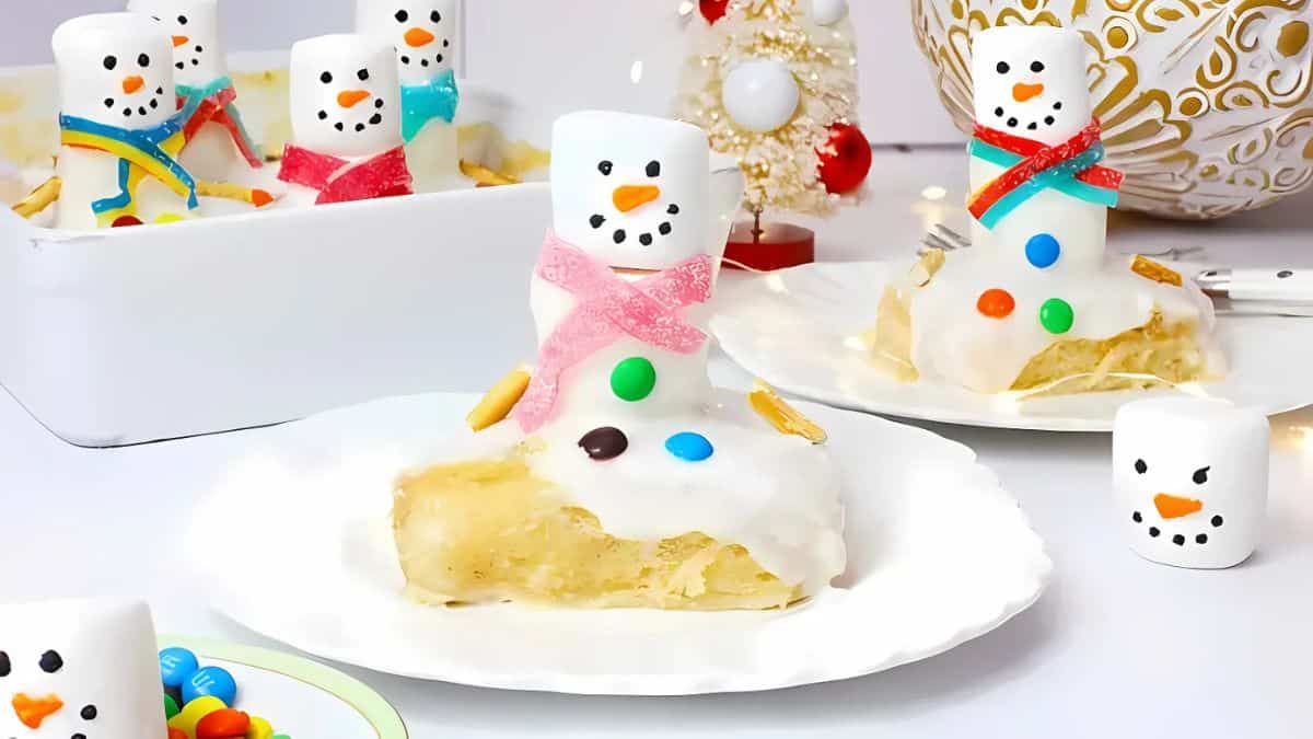 Festive snowman-themed desserts on plates, with marshmallow snowmen topped with colorful candies and pink or red scarves, placed on icing-covered pastries. A small Christmas tree and decorative elements are in the background.