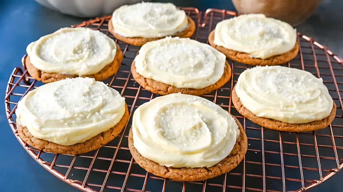 Seven cookies topped with white frosting and sugar sprinkles sit on a round, copper wire cooling rack. The cookies are arranged neatly and displayed on a blue surface.