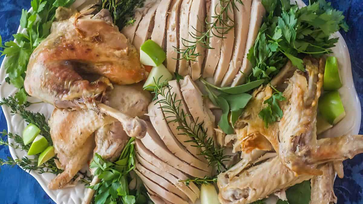 A platter with sliced roasted turkey surrounded by sprigs of rosemary, parsley, and apple wedges. The turkey is golden-brown with both white and dark meat visible. The plate is set against a dark blue background.