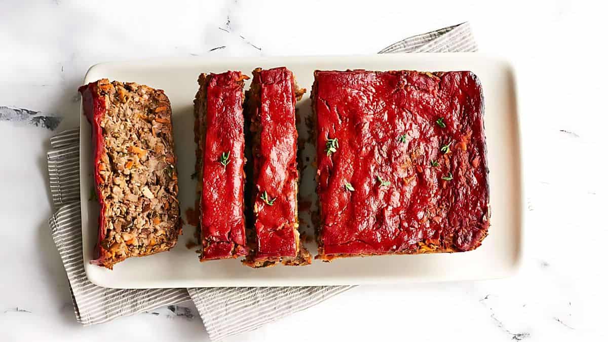 A rectangular, glazed meatloaf sliced on a white rectangular plate, garnished with fresh herbs. The plate rests on a striped cloth over a marble surface.