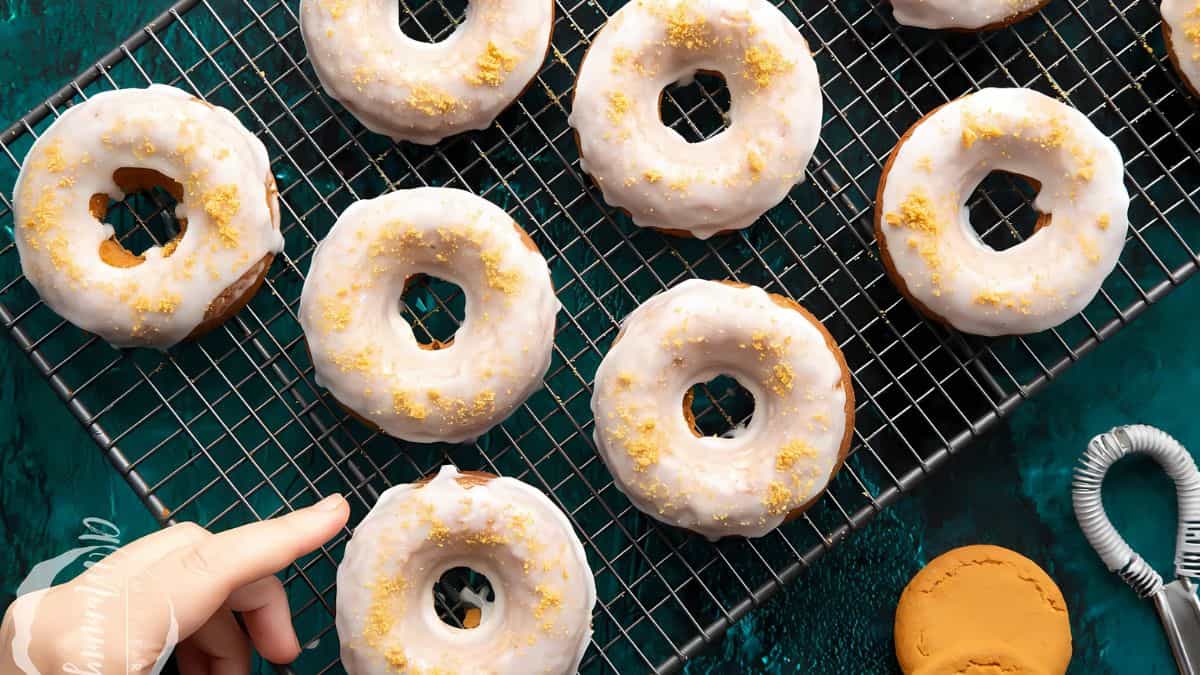 A hand reaching for a glazed donut on a cooling rack, surrounded by other donuts. Each donut is covered with white icing and sprinkled with crumbs. A green background and various baking tools are visible nearby.