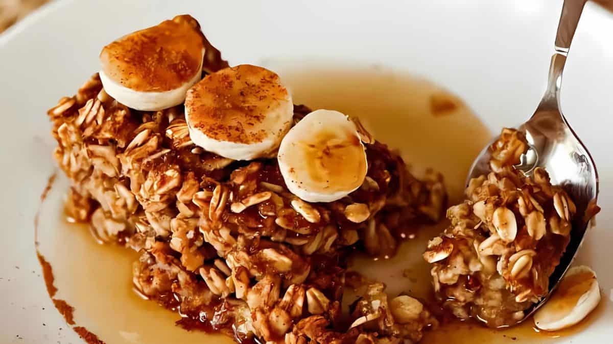 A close-up of a serving of baked oatmeal with banana slices on top, drizzled with syrup. A spoon is scooping a portion from the dish, showcasing the texture of the oats and syrup.