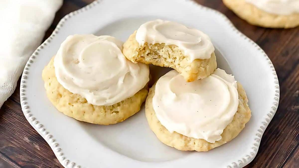 A plate with three frosted cookies. One cookie has a bite taken out of it, revealing a crumbly interior. The cookies are on a decorative white plate, set on a wooden table.