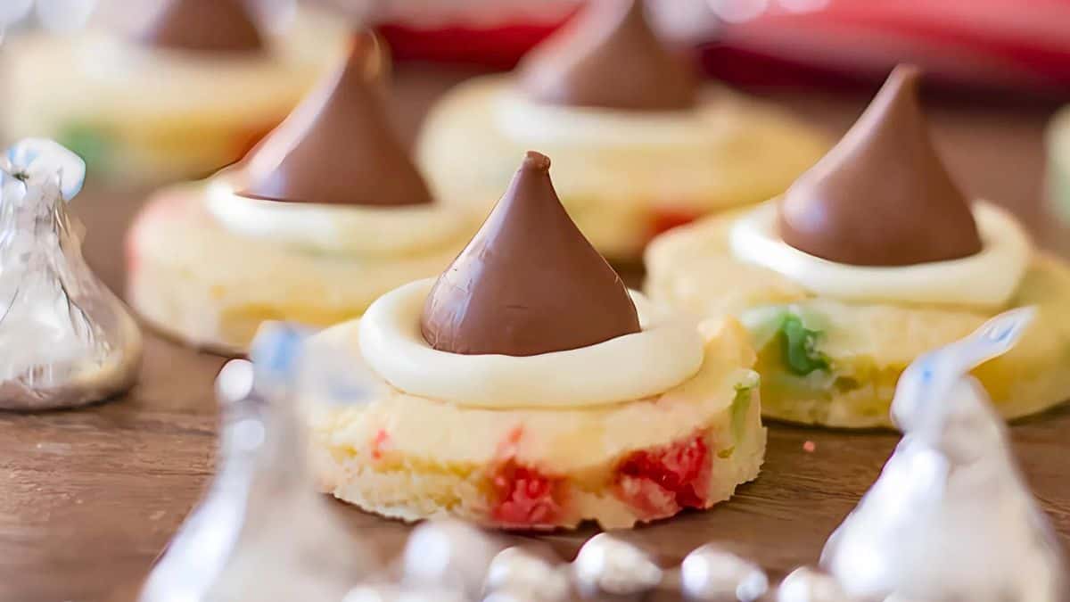 Cookies with colorful bits are topped with chocolate kisses and a swirl of frosting. Silver foil-wrapped chocolates are scattered around.