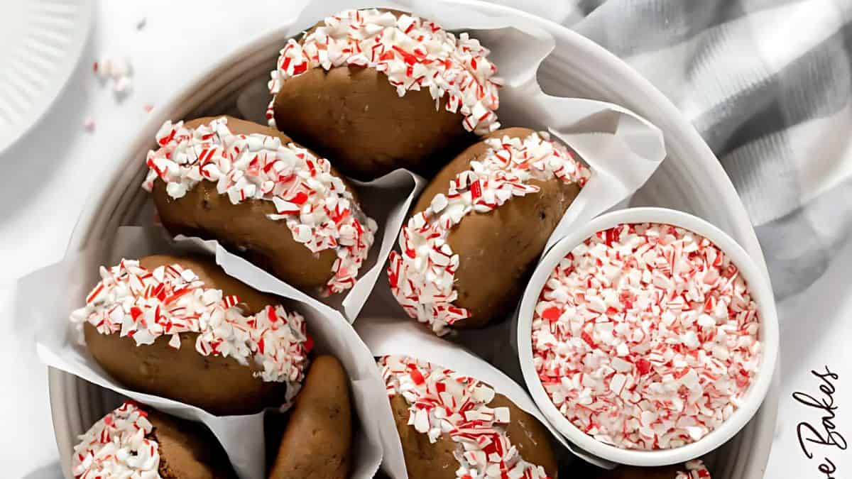 A bowl of chocolate cookies with creamy fillings, each half-dipped in crushed peppermint candies. A small bowl of extra crushed peppermint is nearby. Cookies are neatly arranged in white parchment paper.