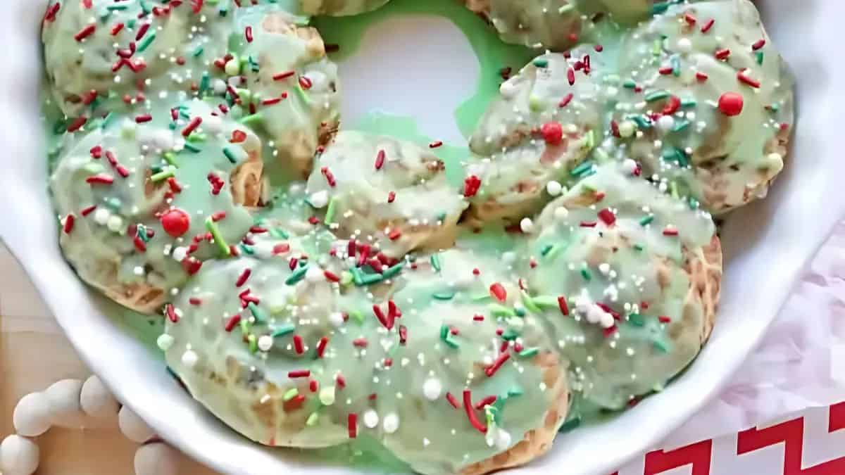 A festive cake wreath decorated with green icing, colorful sprinkles, and red and white candy beads, displayed on a white plate.