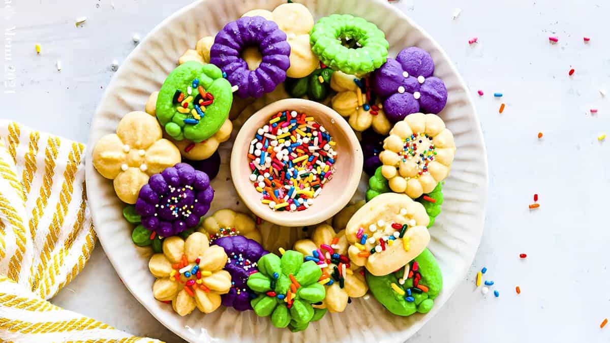 A plate of colorful flower-shaped cookies in shades of purple, green, and yellow, surrounding a small bowl of rainbow sprinkles. A striped yellow napkin is partially visible on the side.