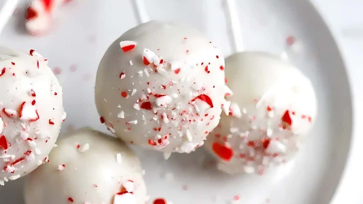 Close-up of three cake pops covered in white chocolate and sprinkled with crushed red and white peppermint candy, placed on a white plate.