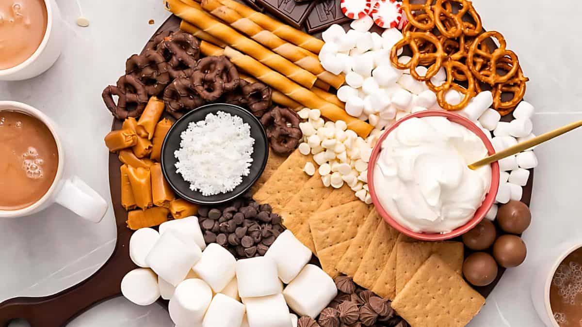 A dessert platter with whipped cream, marshmallows, graham crackers, pretzels, chocolate chips, wafer sticks, caramel candies, chocolate-covered pretzels, and peppermint. Two mugs of hot chocolate are beside the platter.
