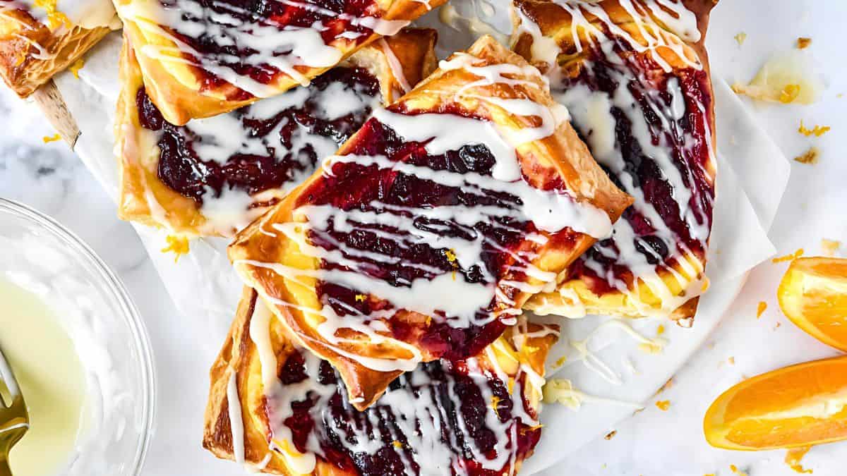 A stack of pastries topped with red berry filling and drizzled with white icing is placed on a white surface. Slices of orange lay beside the pastries, adding a pop of color to the scene.