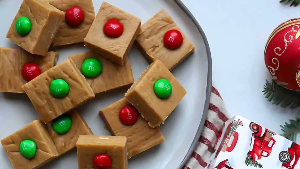 A plate of peanut butter fudge squares topped with red and green candies. The plate is surrounded by a festive Christmas ornament and a gift box with a red truck design.