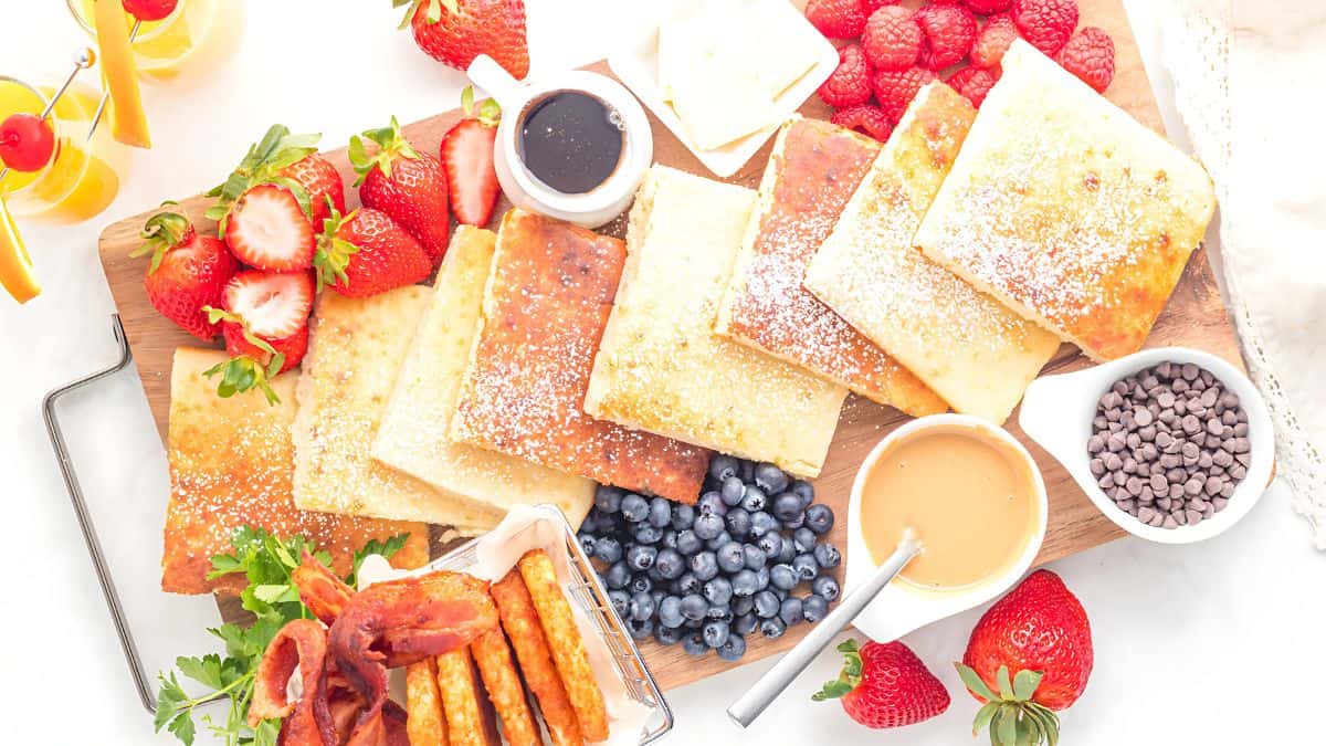A vibrant breakfast spread features pancakes dusted with powdered sugar, strawberries, raspberries, blueberries, crispy bacon, hash browns, chocolate chips, peanut butter, syrup, and orange juice in glasses with straws.