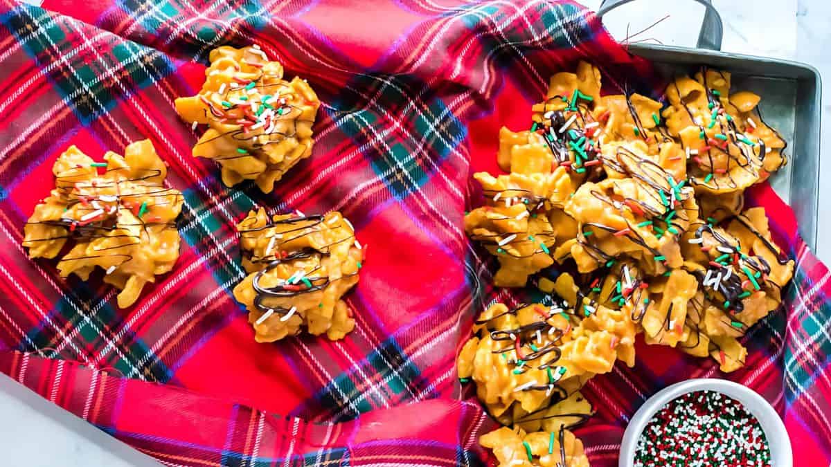 Crunchy candy clusters with chocolate drizzle and colorful sprinkles are displayed on a red plaid cloth, with more clusters in a tray. A small bowl of sprinkles is nearby.