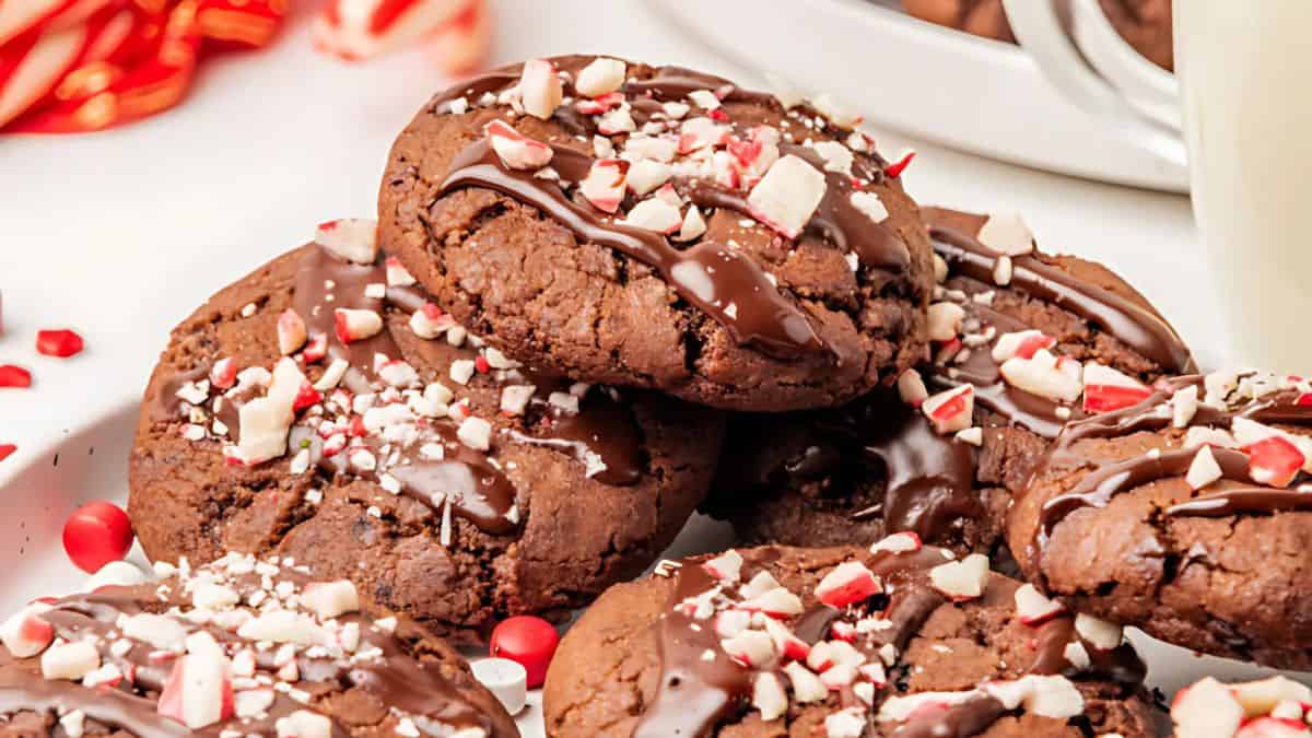 Close-up of a stack of chocolate cookies topped with melted chocolate and crushed candy canes. The cookies are on a white surface, surrounded by red decorations, creating a festive and inviting scene.