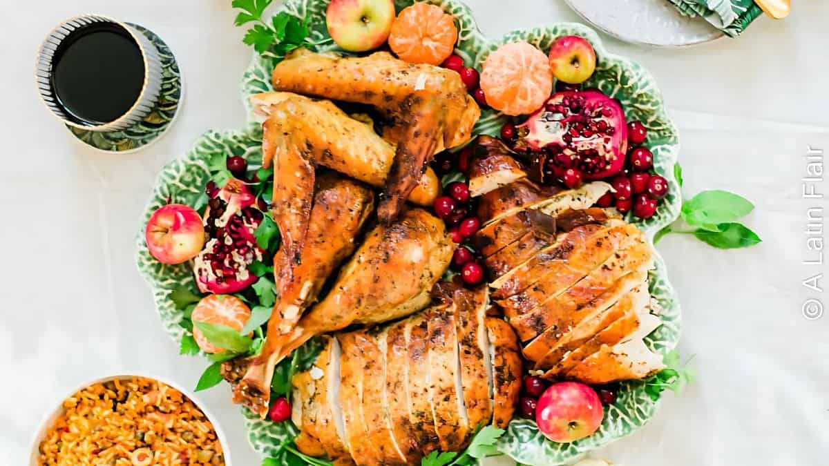 A beautifully arranged Thanksgiving platter featuring roasted turkey pieces, garnished with pomegranate seeds, cranberries, apples, and clementines, next to a glass of wine and a side dish on a white tablecloth.